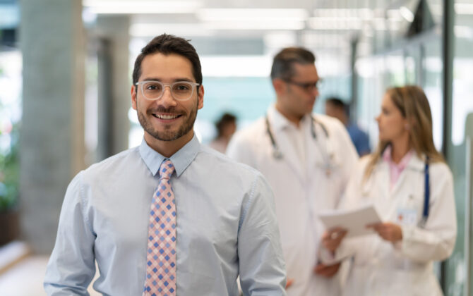 Hospital employee benefits administrator looking at camera smiling while doctors discuss something at background